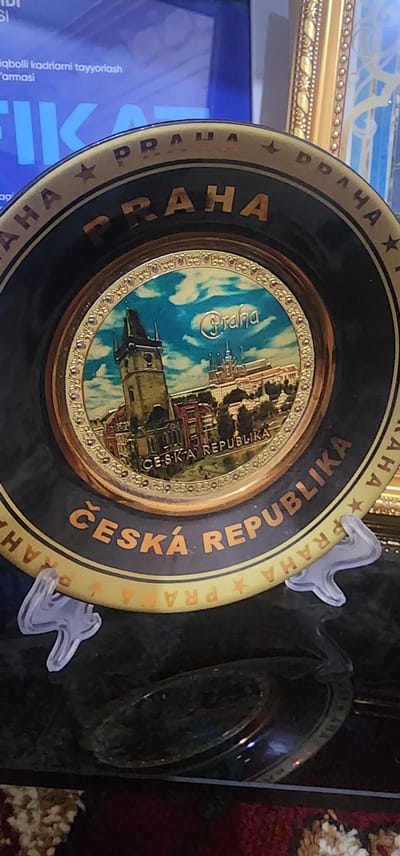 This image depicts a decorative plate featuring a scene of Prague, Czech Republic. The plate is dark with gold accents and has text around the rim. The central image is a painting of Prague Castle and the Charles Bridge. The plate is displayed on a dark surface, possibly a table, with a patterned rug visible in the foreground. There are two clear plastic stands supporting the plate. The background is out of focus, but appears to be a wall with a framed picture and a television screen showing text in an unfamiliar language. The text on the plate is in both Czech and English, reading "Praha" and "ČESKÁ REPUBLIKA" (Czech Republic). There is no indication or visual element within the image that suggests a connection to Jizzakh, Uzbekistan.