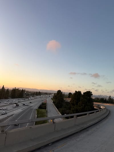This image depicts an elevated view of a multi-lane urban freeway system at dusk, situated in Culver City, United States of America.
The foreground features a grey concrete overpass or ramp barrier, equipped with a metal guardrail containing yellow circular reflectors. The asphalt surface of this structure is visible, showing minor wear.
Below and to the left, a wide freeway with approximately six to eight lanes is visible, divided for opposing traffic flows. This freeway is heavily congested with numerous vehicles, primarily passenger cars and some trucks, moving in both directions. Headlights are visible on vehicles traveling away from the viewer, while the density of vehicles indicates slow movement.
To the right of the main freeway, a narrower, elevated road or ramp appears less utilized, flanked by dense, dark green conifer-like trees. Further right, another section of elevated road curves away from the viewer.
The background consists of rolling hills and a scattered urban landscape with various buildings. The sky transitions from a warm orange-yellow near the horizon on the far left, indicative of a sunset, to a pale blue overhead. A single prominent, diffuse pink cloud is centrally located in the upper sky, with a few smaller, fainter clouds scattered elsewhere. No individual people are discernible within the frame.