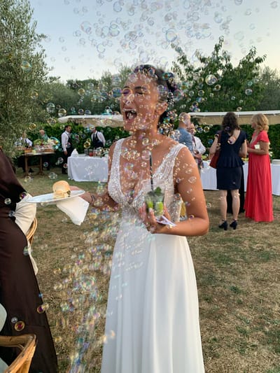 The image depicts an outdoor daytime scene, possibly a wedding reception, with a focus on a woman in a white gown amidst a shower of iridescent soap bubbles. She is in the foreground, her mouth open in a scream or laugh, holding a clear plastic cup with a green beverage and a small white plate with a burger and napkin. Her white dress features a shimmering, beaded neckline. To her left, a person in a dark brown outfit is partially visible, holding a rattan chair. In the background, several individuals are seen near tables, one with white tablecloths. The setting appears to be a garden or courtyard, with dry grass, olive trees, and other greenery. The sky is a pale blue with a hint of yellow as if at dusk. While the image is identified as being in Montepulciano, Italy, there are no specific architectural landmarks or contextual clues within the frame to confirm this location.