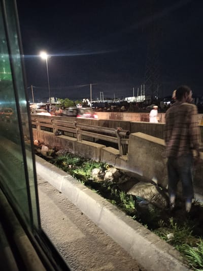 The image, taken at night, shows a roadside scene in Owode-Elede, Nigeria. The view is from inside a vehicle, with the frame of the window visible on the left. A concrete barrier separates the roadside from a bridge or overpass. Trash and vegetation are present along the barrier's base. On the bridge, blurred cars and pedestrians can be seen. A man in a striped shirt and jeans is standing on the roadside, partially obscuring the view. In the background, streetlights and a large utility tower are visible. A building and other structures are discernible in the distance.