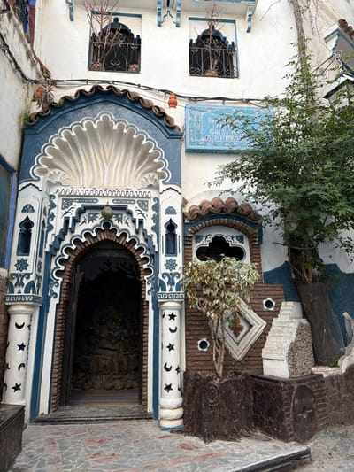 The image depicts the elaborate, blue and white facade of a building, identified as a guesthouse, in Chefchaouen, Morocco. The central feature is a recessed archway entrance with a dark interior. The archway is highly decorative, featuring an ornate scalloped shell motif at the top, intricate geometric and floral patterns in dark blue on a white background, and side pillars with cutouts of stars and crescent moons. Above the archway, two identical rectangular windows are visible, each with dark ornamental iron grilles and small potted plants containing leafless branches.
To the right of the main entrance, a smaller, dark blue tiled window or niche with a quatrefoil-like design is set into a section of reddish-brown brickwork. Below this, various decorative elements are arranged, including a white diamond-shaped relief and several irregular, stepped concrete or stone blocks. A small tree with variegated green and white leaves grows from a brown, carved planter shaped like a tree stump, positioned near the rightmost pillar of the main entrance. Another, larger tree with green foliage is situated further right, growing from a similar dark brown container.
A light blue rectangular plaque, framed with intricate patterns, is mounted on the white wall above the small decorative window. Text on the plaque, partially visible, indicates "Dar S**t**a F*** Guest Ho**" and phone numbers "0613637199/053**". The building's roofline features terracotta tiles and blue trim. The ground in front of the entrance is paved with irregular, reddish-brown and gray stones. No individuals are visible, and no discernible actions are occurring in the scene.