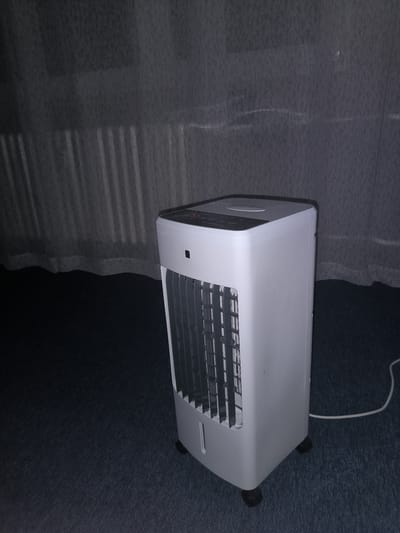 A white portable air cooler stands on a dark blue carpet in front of sheer grey curtains. The air cooler is a rectangular prism with a rounded top, featuring a control panel with buttons and a circular dial. The front of the device has a slatted grille. A white power cord extends from the back of the unit and lies on the carpet, disappearing out of frame. The scene appears to be an indoor setting, possibly a room. There are no people or animals visible in the image. The lighting is dim, with light filtering through the curtains, casting soft shadows. The overall impression is one of a domestic environment, potentially in Kladno, Czechia, though no specific identifying markers of this location are present in the image itself.
