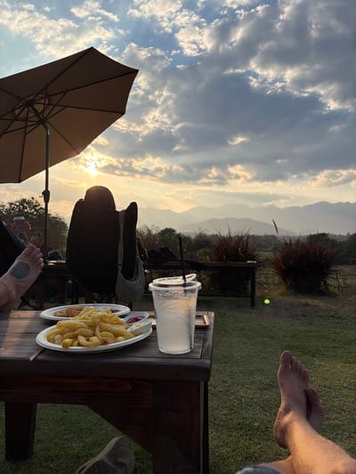 The image captures an outdoor sunset scene in Ban Mae Ping, Thailand. In the immediate foreground, a person's bare left foot with a dark circular tattoo on the top and an ankle bracelet is visible on the left, while their bare right foot and lower leg are crossed and positioned on the right. A dark wooden table occupies the central foreground, featuring a white oval plate mounded with french fries, accompanied by two small white condiment cups containing red and white sauces. Behind this, another white plate holds additional fried food items. A clear plastic cup with a lid, black straw, ice, and a light-colored beverage showing condensation rests next to the plates. To the left, a large, open brown patio umbrella provides shade. A second individual is seated in a dark, high-backed chair, observed from behind, facing the horizon. They have dark hair and a light-colored bag slung over their shoulder. The sun, partially obscured by scattered clouds, is setting directly ahead, emitting golden light and visible crepuscular rays across a distant, multi-layered mountain range. The ground consists of manicured green grass, with several clumps of reddish-brown ornamental grass visible in the mid-ground. Trees and foliage are present on the left and in the distant landscape.
