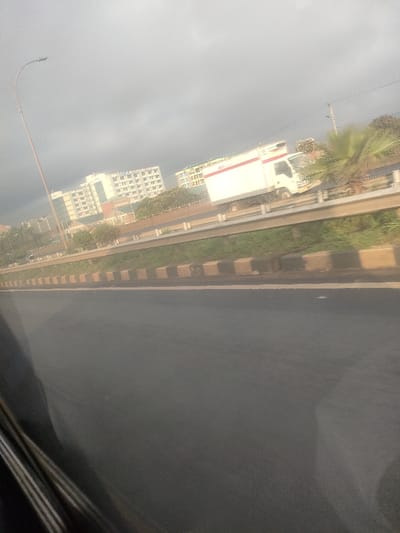The image captures an elevated highway or major road in Theta ward, Kenya, viewed from a moving vehicle. The immediate foreground shows a dark asphalt road surface. To the right, a curb composed of alternating black and light brown concrete blocks borders the roadway. Beyond this curb lies a strip of green grass, followed by a silver metal guardrail. On an elevated section of road behind the guardrail, a white box truck with a red horizontal stripe and a refrigeration unit mounted above the cab is visible, moving towards the right. Multiple light-colored, multi-story buildings are present in the distant background, beyond the elevated road and interspersed vegetation, including shrubs and a large palm-like plant. A tall, slender light pole with a curved arm and luminaire extends vertically on the left side of the frame. The sky is uniformly overcast and light grey. The lower left corner shows a dark, reflective surface consistent with a vehicle's interior trim or window.