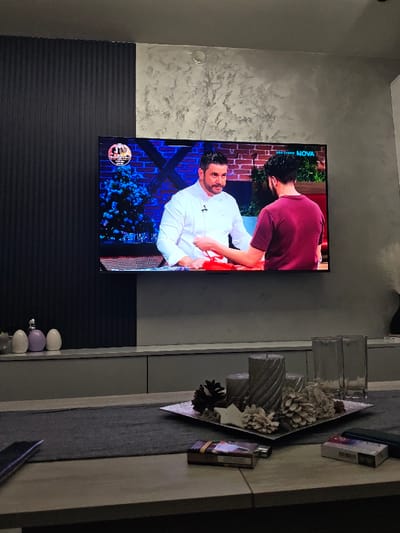 This image depicts a residential interior located in Opaka, Bulgaria. The primary focal point is a large, wall-mounted flat-screen television displaying a culinary program from the Bulgarian channel NOVA. On screen, two adult males are visible: one in a white chef's coat with a dark beard and mustache, engaging with another male in a maroon t-shirt whose back is partially turned to the camera. The television's background features a brick wall, blue accent lighting, and a decorated Christmas tree.

Below the television is a low-profile media console or cabinet in a light gray finish. On the left side of the console, three decorative egg-shaped objects, white and lavender in color, are arranged. The wall above and to the left of the television is divided: the left section is dark with vertical ridging, while the right section is a lighter, textured, possibly stucco or Venetian plaster finish, featuring a round, metallic fixture near the top center.

In the foreground, a wooden table is covered with a dark gray fabric table runner. A decorative centerpiece rests on the runner, comprising a square metallic tray holding three grey, spiraled pillar candles, several natural and bleached pinecones, and a white star-shaped ornament. Adjacent to the centerpiece are two clear, rectangular glass vases. To the right of the centerpiece, two packs of cigarettes are visible on the table; one is open with a metallic lighter nearby, and the other is closed. A dark-colored smartphone is partially visible on the far left edge of the table. The overall environment is dimly lit, suggesting evening or low ambient light conditions.