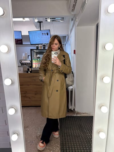 A woman in a trench coat stands inside what appears to be a cafe or coffee shop in Kvarsa, Russia, as reflected in a vanity mirror with multiple lights. She holds a smartphone, possibly taking a picture. The cafe has menu boards displayed, a counter area with drink preparation equipment, and a refrigerated display case stocked with beverages. The floor appears to be tiled or paved. The woman wears black pants and pink and white sneakers.