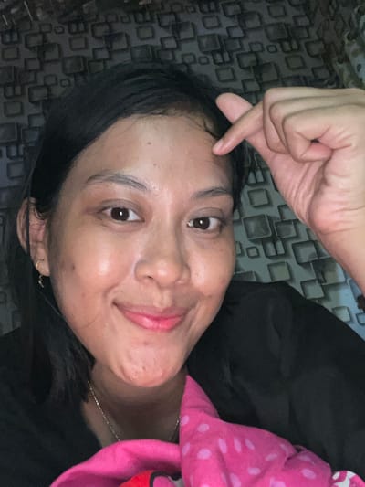 A woman lies on a patterned surface, smiling slightly. Her dark hair frames her face. She wears a dark top. A pink polka-dotted object is near her. She has her hand near her forehead, making a small heart gesture. The image appears to be a selfie taken indoors.