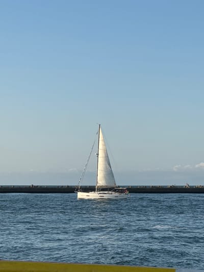 A white sailboat with tan sails navigates the blue water of the Adriatic Sea under a clear sky. A breakwater, constructed of large, dark, angular rocks, stretches horizontally across the middle ground, separating the open sea from the calmer waters near the shore. The sailboat appears to be moving away from the viewer towards the breakwater. The scene does not contain any recognizable landmarks that would specifically place it within the city of Ravenna, Italy.