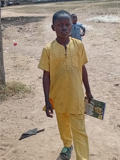 The image depicts an outdoor scene in Abobo, Nigeria, featuring two young boys on sandy, unpaved ground. In the foreground, a boy of approximately 8-10 years old stands centrally, looking directly at the camera. He wears a yellow, short-sleeved tunic with small dark dots and matching trousers, along with green and black open-toed slides. His left hand holds a book titled 'MATHEMATICS' on its cover, which also displays a picture of a boy. To the boy's left on the ground, a dark, crumpled object is visible. In the mid-ground, slightly behind and to the right of the foreground subject, another younger boy, dressed in a light blue long-sleeved garment, stands partially obscured, looking towards the left. The ground consists of dry, light-brown sand with sparse patches of dried vegetation. A small red object, possibly a plastic cup, lies on the ground in the left background. The background shows an open, unpaved area leading to a distant structure or fence line under a bright sky.