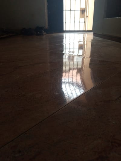 The image presents an interior scene from a low-angle perspective, focusing on a highly polished tiled floor. The floor, composed of dark brown or terracotta-colored tiles, exhibits a reflective surface that prominently mirrors the light entering the room.
In the background, a brightly lit opening, likely a window or a security door, is visible. This opening features a distinct grid pattern of metal bars, which are clearly reflected on the shiny floor. To the left of this opening, a dark door frame is discernible, while a lighter-colored wall or door frame borders the right side.
In the dimly lit left background, several pairs of dark-colored shoes or sandals are arranged near a wall. No people are visible, and no specific activity is taking place, suggesting a quiet, unoccupied interior.
The strong, clear reflections and the significant contrast between the bright light source and the shadowed interior indicate that the photograph was taken during the daytime. The presence of security bars and the style of the tiled flooring are common features in residential settings, aligning with the provided location context of Oke Ogun, Nigeria. The overall impression is one of serene quietude, highlighted by the dramatic play of light and reflection. There is no visible text within the image.