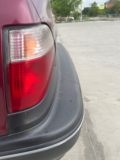 The image shows the rear left portion of a dark red car. The taillight, a combination of clear, amber, and red sections, is prominently featured. The car's black plastic bumper extends across the right side of the frame, showing some dirt and water spots. A thin white stripe runs along the edge of the bumper. The background is an overcast, grey concrete surface, possibly a parking lot or road. To the left, out of focus, are some green trees and what appear to be pedestrian crossing signs with a blue background and white figures. The lighting suggests it might be daytime, possibly overcast. The scene appears to be outdoors in an urban or suburban environment. No people are clearly visible, though the presence of pedestrian signs implies human activity in the area.