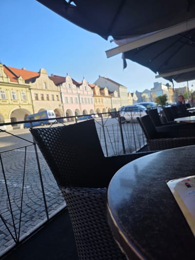 The image presents a street-level view from an outdoor cafe patio in Domažlice, Czechia, specifically looking toward Chodské Square. In the immediate foreground, to the right, a dark, circular outdoor table with a textured surface is partially visible, featuring a light-colored menu item or paper with a red Coca-Cola logo. Adjacent to the table, the back of a dark, woven patio chair is present. A black metal railing with vertical bars runs diagonally across the midground, separating the cafe area from the street. Beyond the railing, the street is paved with grey cobblestones.
In the background, a row of historically styled buildings with prominent gabled roofs and ground-level arched arcades lines the street. From left to right, these include a yellow building, a lighter beige building, a pink building, and a darker yellow/orange building further down the square. A blue van or small bus is visible on the cobblestone street in front of the left-most buildings. Further down the street, several cars are parked or driving, and other outdoor cafe seating areas are visible. Several indistinct figures populate the distant cafe areas and the street. The sky above the buildings is clear and bright blue. The top of the image is partially obscured by a dark awning or overhead structure belonging to the cafe.