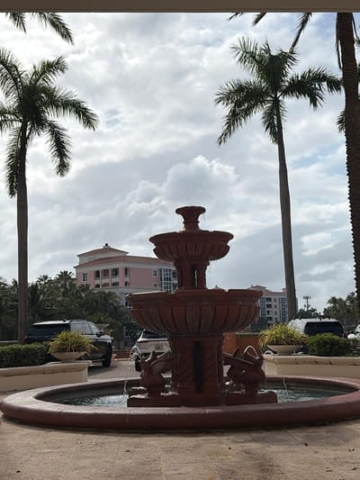 A ground-level, medium-shot photograph features a decorative terra-cotta fountain in the foreground with a Key Biscayne, Florida, United States background. The fountain is multi-tiered, with water flowing from spouts shaped like animal heads into a basin. The basin is bordered by a textured, slightly curved wall. Beyond the fountain, palm trees are visible against a cloudy sky. Several buildings, including a pink building with multi-story levels and a building with numerous windows, are partially visible between the palm trees. Several cars and potted plants, parked along a street are also in the view.