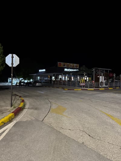 The image depicts a nighttime scene of a McDonald's restaurant located in Gelemenovo, Bulgaria. The sky is dark, indicating a lack of daylight, with artificial illumination provided by the restaurant's signage and ambient lights.
The primary subject is a McDonald's building, single-story with a dark roof structure. Illuminated "McDonald's" lettering and the characteristic "M" golden arches logo are visible on the facade and atop a menu board on the right. Adjacent to the building is an outdoor seating area featuring tables and chairs, partially covered, and equipped with several lamps. To the right of the outdoor seating, a distinctive children's play area, constructed to resemble a train car with red and white elements, is enclosed by a metal fence. A "McDrive" sign, featuring the McDonald's logo, is positioned near the entrance to a drive-thru lane.
In the foreground and midground, an asphalt paved area serves as a roadway or parking lot. The asphalt exhibits wear, with visible cracks throughout its surface. Yellow painted arrows are present on the asphalt, indicating directional flow, along with white painted lines. A curb, painted in alternating red and yellow segments, lines the left edge of the paved area, with some vegetation growing from between the curb and the ground. A white octagonal stop sign, mounted on a dark pole, is prominently situated in the left foreground.
Several vehicles are present in the vicinity of the restaurant. A dark-colored car is partially visible behind the stop sign on the left. Further toward the restaurant, another dark-colored car and a light grey minivan or SUV are positioned. The headlights of a vehicle are faintly visible in the far left background. No individuals are clearly discernible within the frame. Trees and other foliage are visible in the background, appearing dark against the night.