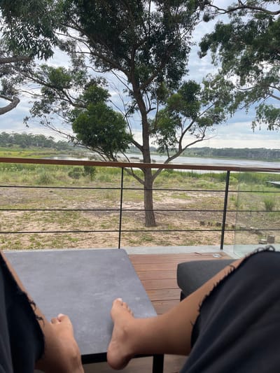 This image depicts a first-person perspective from a private outdoor deck or balcony in La Barra, Uruguay. In the immediate foreground, the bare feet and lower legs of an individual wearing dark, torn-hem shorts rest on or near a flat, rectangular, grey outdoor lounge table. To the right, part of a dark grey upholstered cushion is visible. The floor is composed of wooden decking leading to a modern railing system. This railing features a light brown wooden top rail, black vertical posts, multiple horizontal black cables, and a clear glass panel on the right. Beyond the railing, a sparsely vegetated sandy area extends, leading to a body of water, likely a lagoon or river, bordered by reeds and green foliage. Several trees with green leaves are present, with one prominent tree centered in the midground. The distant background shows a treeline under an overcast sky with scattered grey clouds.