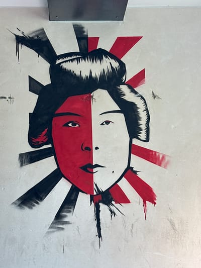 This media file is a photograph of a mural on a wall. The mural depicts two faces, one above the other, in a stylized manner. The faces are split horizontally, with the top half of one face and the bottom half of another, creating a symmetrical and somewhat disorienting effect. Both faces appear to be of people with dark hair styled in a traditional Japanese manner, and their eyes are open and looking in different directions. The mural utilizes a limited color palette of red, black, and white, with red radiating lines emanating from behind the faces, reminiscent of a rising sun motif. A small, silhouetted bird is visible in the upper left quadrant of the mural. The wall itself appears to be a textured, light-colored surface, possibly concrete or stucco. To the left of the mural, a dark metal object, likely part of a building's structure or a fixture, is partially visible. The overall impression is one of bold graphic design and perhaps a commentary on duality or perspective. The location context indicates this mural is in Bonn, Germany. There is no visible text within the image.