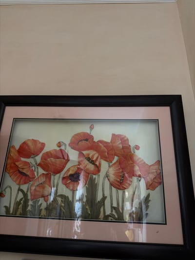 The media file is a photograph of a framed artwork of red poppies hanging on a wall. The artwork is framed with a black outer frame and a light pink mat. The poppies are depicted in various stages of bloom, with some fully open and others in bud. The background of the artwork is a light, neutral color, and there are hints of green foliage. The wall the artwork hangs on appears to be a light, neutral color, possibly beige or cream. There is a white baseboard or molding visible to the left of the artwork. The lighting in the image suggests it was taken indoors, possibly during the daytime due to the reflections on the glass of the frame, which hint at ambient light from a window or room. There is no visible text in the image. The scene is domestic or interior, with the focus being on the framed artwork. The image is cropped to emphasize the artwork and the wall it is on.