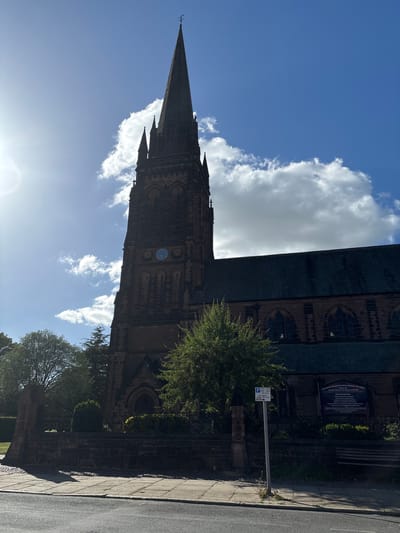The image captures a view of The Parish Church of St Mary Without-the-Walls, located in Chester, United Kingdom. The church's steeple rises prominently against a blue sky dotted with white clouds. Architectural details of the stone structure, including arched windows and decorative elements, are visible. A clock is visible on the tower. Alongside the church, a section of the sidewalk and a nearby street can be seen. A parking sign and a notice board titled "The Parish Church of St Mary Without-the-Walls Ancient Parish of St Mary-on-the-Hill Church Services" stand adjacent to the building. Trees and greenery are present around the church.