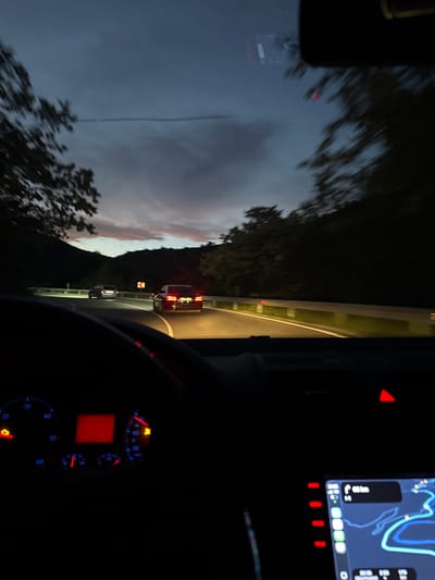 The image shows a view from inside a car driving on a road near Kremen, Bulgaria. A red car is ahead, and a white car is further ahead. The sky is a twilight color, with trees lining the road. The dashboard of the car is visible, including the navigation system and speedometer. The road is curved.