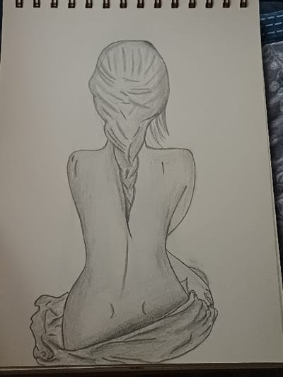 The image displays a photograph of a pencil drawing on a white, spiral-bound sketchbook. The drawing depicts the back of a seated female figure, rendered from the shoulders to the upper thighs. The figure's head is seen from behind, with hair styled and a single braid extending down the center of her back. The back is gently curved, showing the contours of the spine and a slender waist. A draped piece of fabric, possibly a sheet, covers the figure from the lower back downwards, gathered in folds around her hips and thighs. The drawing utilizes shading to create depth and form. The sketchbook features a black metal spiral binding along its top edge. The sketchbook rests on an unidentified flat surface, with a dark horizontal line visible below its bottom edge, and a section of blue and white patterned fabric, possibly upholstery or a blanket, visible to the right of the sketchbook. No discernible features within the drawing or the photographic context indicate a specific physical location in the city of 서대문구, South Korea, or any other geographic area.