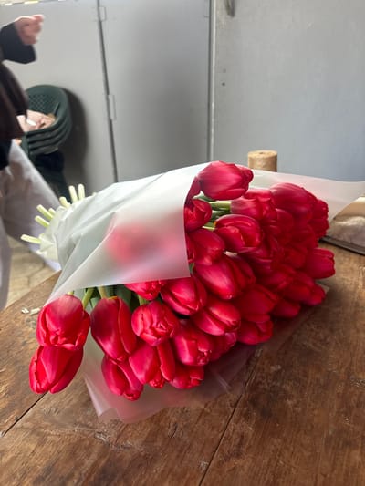 A large bouquet of numerous closed red tulip blossoms, wrapped in translucent white paper and clear plastic film, is centrally positioned on a rustic, dark brown wooden tabletop in an indoor location within Trudobelikovskiy, Russia. In the upper-left background, a partially visible individual, dressed in light-colored trousers and a dark upper garment, has hands active above a stack of green plastic chairs. The background wall is light grey, featuring two distinct vertical panels, likely doors. On the table in the upper-right, a brown twine spool stands near a segment of light-colored fabric. The overall scene suggests a floral preparation or storage area.