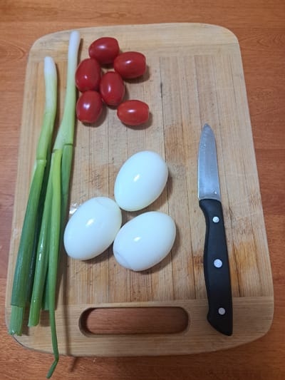An overhead, medium shot captures a wooden cutting board on a wood-grain table. The cutting board is positioned slightly to the left of the frame's center and displays several food items and a knife. From the top left corner moving clockwise, there are four green onions, their white bases visible. To the right of the onions are six small, red cherry tomatoes, clustered together. Below the tomatoes and slightly to the left are three peeled, hard-boiled eggs. To the right of the eggs and along the right edge of the cutting board lies a paring knife with a black handle. The background consists of a wooden table with a prominent wood grain pattern. No people are visible in the image. The scene appears to be a preparation for a meal. The specific location in the City of Moncton, Canada, is not discernible from the visual information provided in the image.
