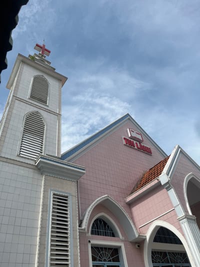 A low-angle shot captures a building with distinct architectural features against a cloudy sky. The building has a white tower with a red cross on top. Adjacent to the tower, a pink section is marked with red signage. The building design includes arched windows and horizontal louvers, detailed with white and blue trim. The photograph's perspective emphasizes the height and structural elements of the architecture.