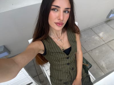 A young adult woman with long dark hair and light skin, wearing a green pinstripe vest over a black top and a green purse with a gold chain, is taking a selfie outdoors. She is sitting on a white chair on a patio with gray tiled flooring. The woman is wearing necklaces, one with a cross pendant and another with a rectangular pendant. The background includes a light gray wall and a light fixture mounted on it. The scene is set in Hallandale Beach, Florida, United States of America.