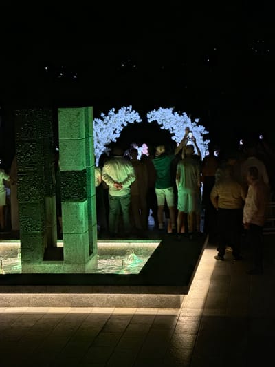 The image depicts a gathering of people at night, illuminated by a combination of artificial lighting and distant light sources. In the foreground, there is a modern water feature with two large, textured, green-tinted stone pillars. The water in the feature is illuminated with green lights. The pillars appear to have intricate carvings. Behind the water feature, a group of people are standing, some holding up their phones, presumably to take pictures or videos. Above the crowd, a floral archway, possibly made of white flowers, is visible. The background is dark, with scattered bright points of light that could be distant city lights or stars. The ground in front of the water feature is paved with rectangular tiles. The overall impression is an outdoor event taking place in the evening. The location is specified as Razliv, Bulgaria.