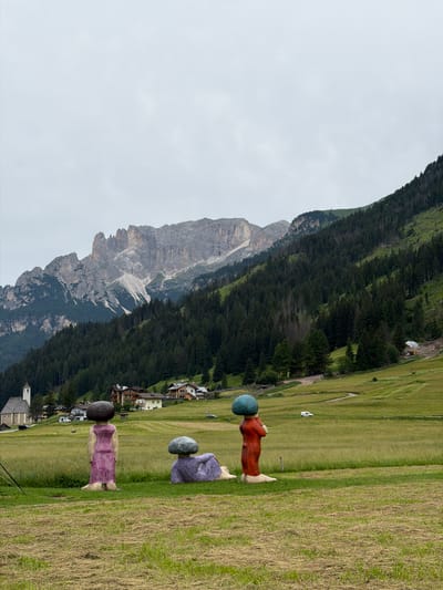 This image depicts a scenic mountainous landscape in Campitello di Fassa, Italy. In the foreground, three abstract human-like sculptures stand on a grassy field. The leftmost sculpture is a figure in a purple dress with a dark head. The middle sculpture is a figure in a red outfit with a teal head. The rightmost sculpture is in a seated position and is purple.
The background features a vast, green, rolling hillside, dotted with trees and a few buildings, suggesting a small village or resort area. Beyond the hillside, a rugged, grey mountain range dominates the scene, with a large, rocky face showing signs of erosion and scree slopes. The sky is overcast and a light grey, indicating a cloudy day.
The composition is vertical, and the image is rotated, making the sculptures appear to be standing on a vertical plane rather than a horizontal one. The lighting suggests it is daytime, but the cloudy weather creates a diffused light. The overall impression is one of a tranquil, natural setting with artistic elements integrated into the landscape. There is no visible text in the image.