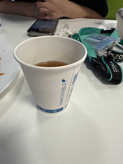 This is a close-up, eye-level shot of a white, compostable paper cup filled with a light brown liquid, likely tea, resting on a white surface. The cup has blue text that reads "COMPOSTABLE" vertically down its side, along with a blue leaf logo and repeating blue "alpha" logos near the bottom rim. To the right of the cup is a teal pouch containing blue and white items, possibly cards or stickers, with a keychain attached. The keychain has black and teal straps with white text. A smartphone is visible in the background, lying on the white surface. Partially visible at the top are the hands of two individuals, suggesting they are sitting at a table. The overall environment appears to be an indoor setting with a neutral lighting. The image does not provide any specific information to determine its location in Chociule, Poland.