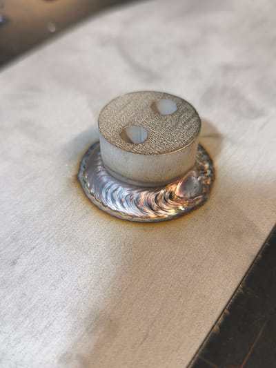 A close-up, high-angle shot shows a metal cylinder with two small holes on top, welded to a metallic surface. The weld forms a ring around the base of the cylinder, exhibiting iridescent colors ranging from blue to gold, indicating heat exposure. The surface beneath appears to be a brushed or textured metal. The background is out of focus, with a suggestion of a darker surface on the right side. No people or specific city landmarks are visible to determine the location in Bad Honnef, Germany. The scene appears to be an indoor workshop or laboratory setting.