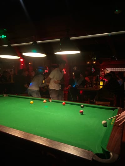 A bar scene includes a pool table covered in green felt, with several pool balls scattered on its surface. A hand holding a cue stick is poised to strike the white ball. People stand around the table, some engaged in conversation. Overhead, three bright lamps illuminate the area, casting light on the scene below. The background features the bar’s interior, with various patrons and decor. The overall ambiance is dim and lively.