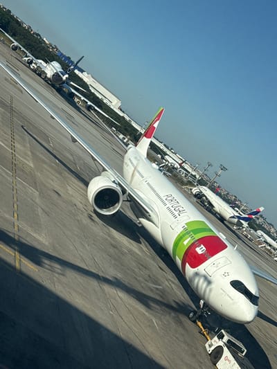 The image depicts an airport tarmac scene in Guarulhos, Brazil, under a clear blue sky. A large white TAP Air Portugal Airbus A330-900, named "INFANTE D. HENRIQUE," dominates the foreground, angled diagonally from the lower left to the upper right. Its fuselage features prominent "PORTUGAL" text and a distinctive green, white, and red livery. A white ground tug, branded "WFS," is attached to the nose gear of the TAP aircraft. In the mid-ground to the left, a Lufthansa Boeing 747-8 is parked, visible with its blue tail logo. Further in the background to the right, a white LATAM aircraft, displaying its red and blue tail livery, is parked near other indistinct airplanes and airport infrastructure, including tall light poles. The tarmac is concrete, marked with yellow lines. Strong shadows are cast across the tarmac from the aircraft and other structures. Distant urban development and green foliage are visible on the horizon. The photograph is taken from an elevated perspective, likely a terminal window, with a slight counter-clockwise tilt.