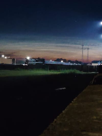The image captures an industrial landscape at dusk or night within the Краснадворскі сельскі Савет, Belarus. The sky transitions from a deep blue at the top to lighter hues of orange and purple near the horizon, suggesting the sun has recently set. In the mid-ground, a series of industrial structures, including what appear to be pipes or conduits, are illuminated by artificial lights, some of which are bright and create lens flares. Two tall radio towers or similar structures are visible in the distance. The foreground is dominated by a dark, grassy area, with a concrete or stone embankment on the right side of the frame. The overall scene conveys a sense of industrial activity against a darkening natural backdrop.