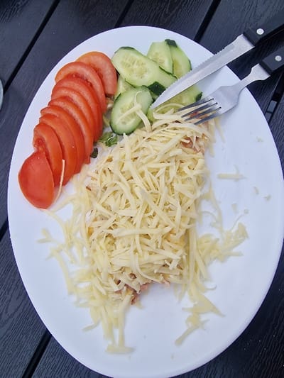 The image depicts a white oval plate, viewed from above, resting on a dark, slatted table surface in the city of Bicaz, Romania. The plate holds a meal consisting of several food items. On the left side, approximately nine uniformly sliced red tomatoes are arranged. In the upper central area, five to six slices of green cucumber are visible. The majority of the plate's surface, particularly the lower and central portions, is covered with a substantial quantity of light yellow shredded cheese, beneath which an underlying, partially obscured food item (likely a main dish) can be seen. A silver serrated knife with a black handle and a silver four-tine fork with a black handle are positioned diagonally across the upper right section of the plate, resting on the cucumber and shredded cheese. The dark, textured table surface, possibly wood or composite material, extends around the plate, suggesting an outdoor or patio setting.