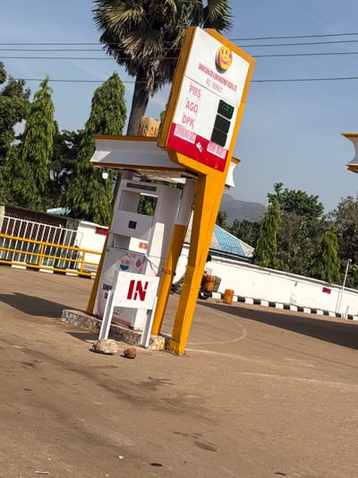 The image displays a static outdoor scene of a fuel station in Tataura, Nigeria, under a clear blue sky. A prominent yellow-framed sign, angled upwards and to the right, is central. It displays "S.E.O & SONS (SUNDAY OLORUNTOBA & SON MANAGEMENT NIGERIA LTD. RC: 1099521)" at the top. Below this, "PMS", "AGO", "DPK", and "COOKING GAS" are listed vertically, each with a blank black rectangular display panel where prices would be. The sign also features "MINI MART" at its base.
Below the sign, two white fuel dispensers are situated under a white canopy supported by yellow pillars. One dispenser is marked "S.E. ORI" and displays a red "NO SMOKING" warning sign. A white sign with red "IN" lettering stands on the asphalt ground in front of the dispensers, stabilized by two small rocks.
The immediate background on the left includes a yellow metal fence and gate. To the right, a white wall with black and white striped curb markings separates the foreground from a background area where a dark-colored motorcycle with an attached storage box and a yellow jerry can are visible. A building with a blue corrugated roof is partially obscured behind this wall. The distant background features lush green trees, including a tall palm tree and several conifers, with mountains visible on the horizon under a clear blue sky traversed by power lines. No individuals are present, and no active interactions are depicted.