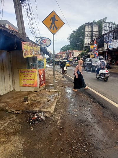 The image captures a bustling street scene in Cimaung, Indonesia, under an overcast sky. The ground appears wet and muddy, suggesting recent rain or damp conditions.

In the foreground to the left, there is a small, white-painted wooden stall. Two banners are displayed on the stall; one with images of food items and text partially obscured, and another yellow banner with "Pisang Kumbung," "Varian Rasa," "Mulai 10K," "- Indomie Rebus," "- Kopi," and "Sedia Masakan" (meaning "Banana Kumbung," "Flavor Variety," "Starting from 10K," "Boiled Indomie," "Coffee," and "Serving cooked food"). Litter and debris are visible on the muddy ground in front of the stall.

A yellow, diamond-shaped pedestrian crossing sign stands prominently on a pole in the mid-ground, marking a pedestrian area. Behind this sign, a blue oval sign reads "PLANET BAN," with smaller text "PUSAT SERVIS" and "AKI MOTOR" (indicating a service center for tires and batteries).

To the right of the sign, a woman wearing a light brown hijab and a long black skirt is seen walking across the street, her back mostly to the viewer. She is stepping near the white line separating the shoulder from the main road. Simultaneously, a person on a white scooter, wearing a helmet and a backpack, is riding past her on the road. A black car is also visible behind the scooter.

The background reveals a row of various shops and buildings lining the street, suggesting a commercial area. Large billboards or structures are visible on a hillside further back, which is covered with dense green foliage. The overall setting depicts a typical, busy urban or semi-urban street environment during daytime. A partially visible red banner with "ERPAI" is also seen on a building on the right.
