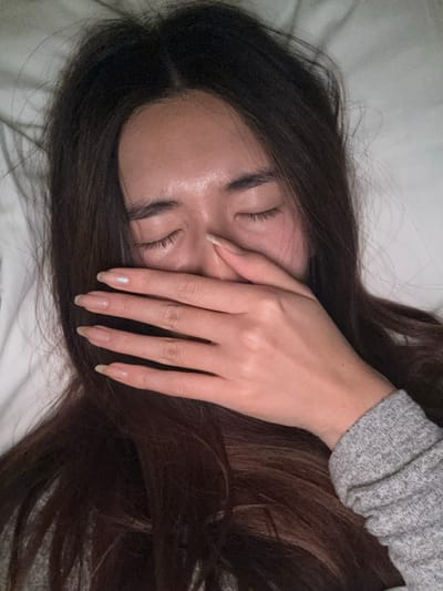 A close-up, top-down view depicts an individual, likely a female, with long dark brown hair, lying down in an indoor setting identified as Le Havre, France. The eyes are closed, and the right hand is positioned over the mouth and lower nose, with the thumb and index finger contacting the bridge of the nose. The individual's skin on the forehead exhibits a slight sheen. Fingernails are long and oval-shaped, with a pale iridescent finish visible on the ring finger. A gray, ribbed long-sleeved garment is visible on the lower right arm. The background consists of white fabric, indicative of bedding, such as a pillow or sheet. The specific location within Le Havre, France, is not visually discernible from the image, which focuses exclusively on the individual and their immediate personal space.