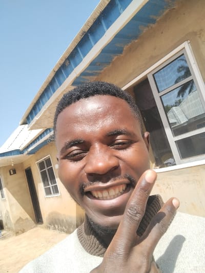 A dark-skinned male subject, approximately 20-35 years old, with short dark hair, a mustache, and a short beard, is prominently featured in a close-up, selfie-style photograph taken in the city of Kaninkon, Nigeria. He is smiling broadly, revealing his teeth, with his eyes squinted, likely due to bright sunlight. He wears a light-colored, possibly off-white or light grey, sweater with a brown turtleneck-style collar. His right hand is raised near his face, forming a 'V' gesture (peace sign) with the index and middle fingers, partially obscuring his chin and mouth.
The background shows a building under clear, bright daylight. The building features light tan or beige, rough-textured stucco walls. A blue corrugated metal roof with white eaves/fascia board is visible at the top. Two white-framed windows are present: one on the upper right with reflective panes showing reflections of what appears to be a palm tree and another building, and a smaller one on the lower left with a grid-like pane structure. A dark-colored door is partially visible on the far left side of the building. Strong sunlight casts sharp shadows from the subject and the building elements. The sky in the upper left portion of the image is clear and light blue. The ground visible on the lower left is light-colored, likely concrete or packed earth.