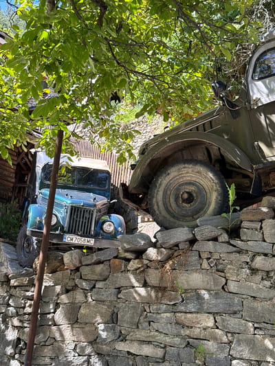 Two vintage trucks are parked on a rocky incline, partially overgrown with vegetation, in Shiroka Laka, Bulgaria. The truck in the foreground is a dark blue, possibly green, open-top vehicle with a visible license plate reading "CM 7058 MC". It is positioned at an angle against a stone wall, with its front wheels resting on larger rocks. A rusty metal pole stands vertically to the left of this truck. Behind and to the right, a larger olive green truck is visible, its front fender and large tire prominent. This truck is also parked on the rocky slope. The environment is lush with green foliage overhead, casting dappled shadows, and a backdrop of stone structures and greenery. The scene appears to be an outdoor setting, likely a rural or historical area, given the style of the trucks and the stone construction.
