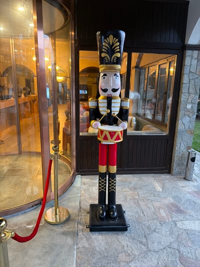 The image depicts the exterior entrance of a building, identified as being in the city of Баня, Bulgaria, during evening hours. A prominent, large, wooden nutcracker figure stands centrally on the paved ground. The nutcracker is dressed as a drummer, featuring a black shako hat with gold ornamentation, a white face with a black mustache and blue eyes, a black jacket with gold and white trim, red trousers with gold stripes, and black boots with gold lacing details. It holds a red and gold drum with white drumsticks.
To the left of the nutcracker is a brass-framed revolving glass door, partially demarcated by a red velvet rope suspended between two brass stanchions. Through the glass of the revolving door, an illuminated interior lobby with warm lighting, a reception desk, and light-colored walls is visible. To the right of the nutcracker are large windows with dark frames, situated above dark wooden paneling. Through these windows, a well-lit interior space with seating, including sofas and chairs, is visible. Adjacent to the windows, a wall is clad in light grey, irregular stone masonry. A silver-colored cylindrical outdoor ash bin rests against this stone wall. The ground in the foreground is paved with irregular, light-toned stone tiles. No individuals are visible in the scene.