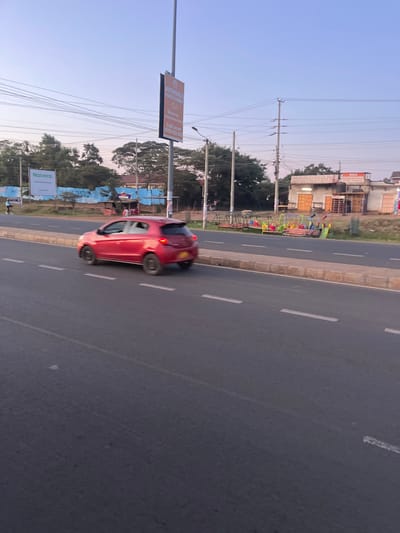 The scene depicts an urban roadside in Biashara ward, Kenya, under a clear sky exhibiting light blue and faint pinkish hues. A multi-lane asphalt road with white dashed lane markings occupies the foreground, separated by a raised concrete median barrier from the opposing carriageway. A red five-door hatchback, consistent with a Mitsubishi Mirage model, is travelling away from the camera in the lane adjacent to the median; its rear wheels are blurred, indicating motion, and a yellow license plate is visible.

To the left of the road, behind the median, a large white billboard featuring "Naivera" text is positioned in front of a blue barrier and dense green foliage. A lone individual is visible walking or cycling in this distant area.

To the right of the road, beyond the median, a tall pole supports a double-sided billboard advertising "AUTHORISED PRINTS & COPIES," "CJ," "Mpesa," and contact information including a phone number (0790 922 022). Adjacent to this, multiple sets of brightly colored children's playground equipment, including swings and slides, are arranged along the roadside. Further right, a single-story building structure houses several businesses, identified by signs as a "BAR," "PROVISION STORE," and "RYAN PROVISION STORE," featuring multiple closed roll-down shutters. Overhead utility poles with numerous power lines are prominent across the entire scene.