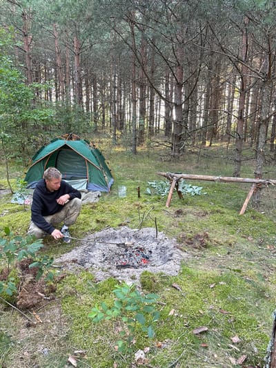 A young man with blond hair is crouching in a forest clearing near a green tent. He is wearing a black hoodie and camouflage pants. He is holding a stick over a campfire that is smoldering with red embers. In the background, there is a wooden structure with some leaves piled next to it. The ground is covered in moss and pine needles. The forest appears to be dense with tall pine trees. The scene suggests a camping trip in a rural area of Belarus.
