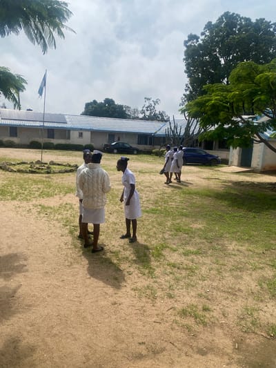 The image depicts an outdoor scene in Dutsen Kato, Nigeria, featuring a group of individuals, likely students, dressed in white uniforms and caps, possibly nurse's uniforms. Four individuals are prominently visible in the foreground, engaged in conversation. Two are standing close together facing away from the camera, while a third stands to their right, looking at them. A fourth individual, wearing a cream-colored jacket over their uniform, stands in the center, facing left.
In the background, a long, light-colored building with a blue metal roof stretches across the frame. Solar panels are visible on the roof. A flag on a pole stands in front of the building. Several trees, including a large one with dense foliage and a smaller one with vibrant green leaves, are present in the scene. Two cars, one black and one blue, are parked near the building. The ground is a mixture of sand and sparse green grass, with a circular stone arrangement visible in the mid-ground. The sky is partly cloudy, suggesting daylight.