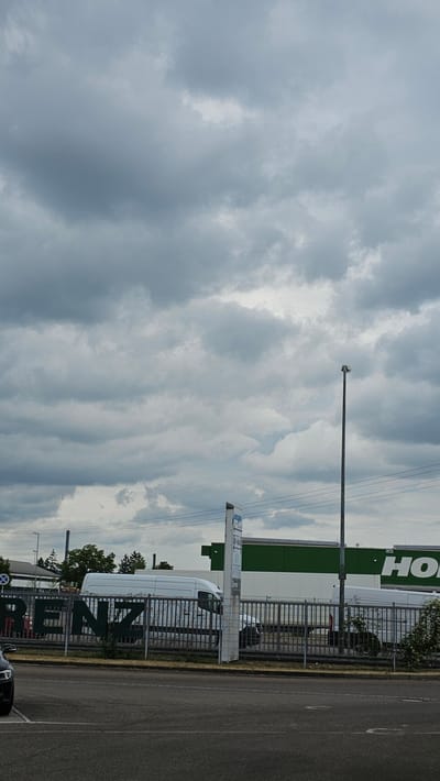 The image depicts a street scene in Neu-Ulm, Germany, under a cloudy sky. In the foreground, a paved road is visible with a black car partially in the frame on the left. Behind the road, a metal fence runs horizontally across the image. Beyond the fence, several white vans are parked or driving. Large, dark green lettering on the fence spells out "RENZ". In the background, a green building with white accents is visible, and further to the right, large white letters on the building appear to spell "HO". A tall, slender street lamp stands on the right side of the image. The overall environment suggests an industrial or commercial area.