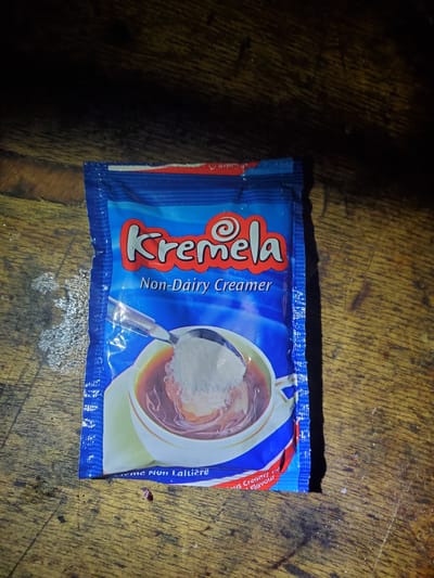 The image shows a single-serve packet of "Kremela Non-Dairy Creamer" placed on a wooden surface. The packet has a blue background with red and white accents. The brand name "Kremela" is prominently displayed in large, stylized white lettering with a red outline. Below the brand name, "Non-Dairy Creamer" is written in smaller white text. A graphic on the packet depicts a white mug filled with a brown liquid, possibly coffee or tea, with a spoon scooping white creamer into it. The packaging also features text in a language that appears to be French: "Crème Non Laitière" which translates to "Non-Dairy Cream." The wooden surface appears aged and textured, with visible grain and some scattered debris. The lighting suggests an indoor setting, possibly with natural light creating shadows. The overall impression is of a product ready for use, possibly in a home or a small shop setting in Barikin Sabon Gidan Kanat, Nigeria.