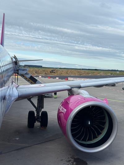 An airplane with pink accents is parked on a tarmac. The aircraft's fuselage, wing, and engine are visible in the foreground. The airplane's tail fin, painted in shades of pink and purple, rises vertically in the upper left corner of the image. A set of stairs, labeled "ALLgate," is attached to the side of the aircraft. The airplane's landing gear with two wheels is visible on the ground. In the background, construction activity is evident with a bulldozer and piles of dirt. Fencing and barriers are also present. The sky above is filled with white, wispy clouds. The scene appears to be at an airport. The provided location is Künersberg, Germany.