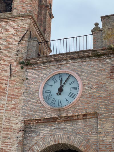 The image shows a close-up of a clock tower in Barbara, Italy. The clock face is circular with Roman numerals and has two hands. The clock tower is made of brick and has a railing around it. The sky in the background is overcast. There are no people visible in the image.