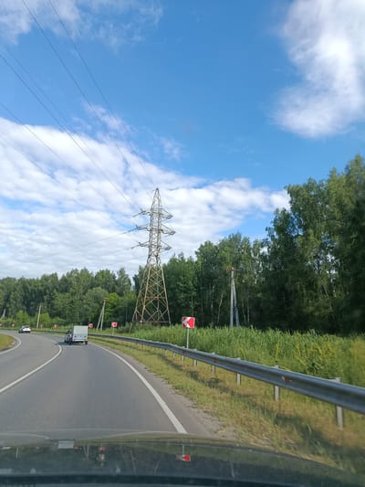 This media file captures a view from a vehicle traveling along a road in Тополиный, Russia. The scene depicts a sunny day with a blue sky dotted with white, fluffy clouds. A large, lattice-style electrical tower stands prominently on the right side of the road, with multiple power lines stretching across the sky.
The road itself is paved and curves to the left. There are two vehicles visible: a white car in the distance on the left lane and a white trailer being towed by another vehicle in the right lane. The road is bordered by a metal guardrail on the right, with tall green grass and trees forming a dense backdrop. Red and white directional signs indicating curves are visible along the roadside. The overall impression is that of a typical summer day in a rural or semi-rural area, with a focus on the infrastructure and natural landscape. The time of day appears to be late morning or early afternoon, given the bright sunlight and clear sky.